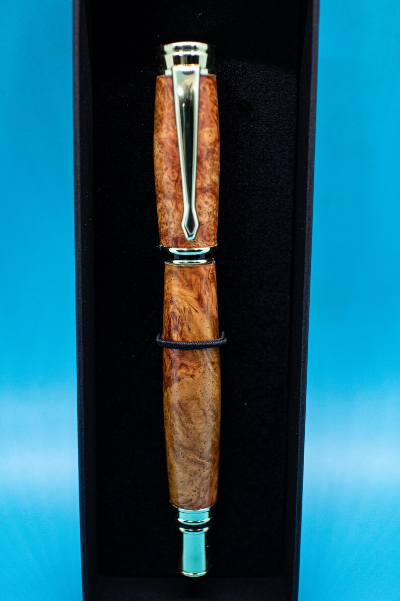 May include: A handcrafted pen with a wood-grain barrel and gold-colored accents, presented in a black box. The pen features a silver clip and a black band. The pen's design showcases a natural wood pattern, adding to its elegant appearance.