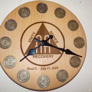 May include: A wooden clock with a black face and hands. The clock is decorated with twelve AA sobriety coins, each with a different number of years of sobriety. The clock also features the AA triangle logo with the words "Unity", "Service", and "Recovery" inscribed on it. The clock is personalized with the name "Brad C." and the date "July 17, 2009".