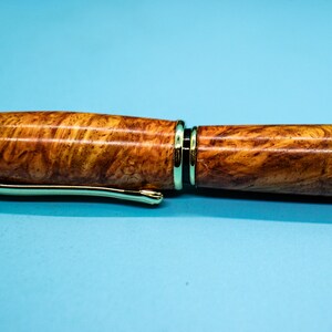 May include: Close-up of a wooden pen with a gold-colored clip and accents. The pen's body has a natural wood grain pattern in shades of brown and yellow. The pen is set against a light blue background.