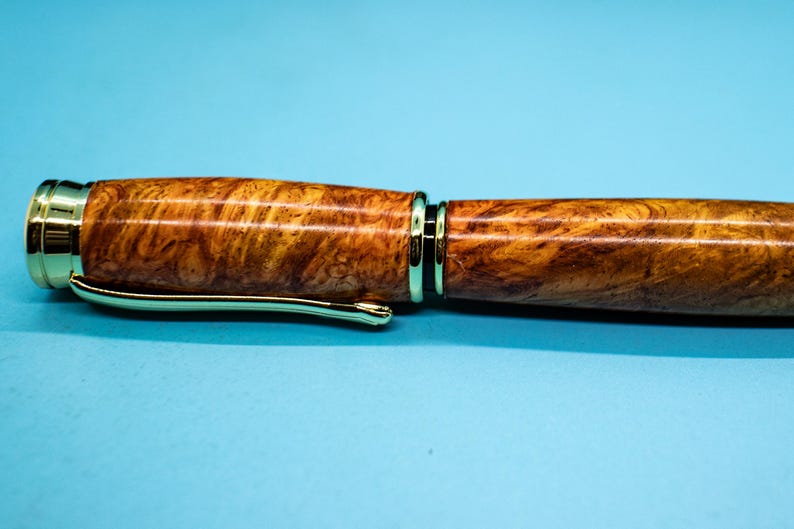 May include: Close-up of a wooden pen with a gold-colored clip and accents. The pen's body has a natural wood grain pattern in shades of brown and yellow. The pen is set against a light blue background.