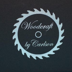 May include: A circular logo featuring a light blue saw blade design with the words "Woodcraft by Carlson" in a script font. The logo is set against a dark background, creating a contrast.