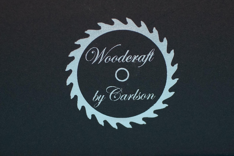 May include: A circular logo featuring a light blue saw blade design with the words "Woodcraft by Carlson" in a script font. The logo is set against a dark background, creating a contrast.