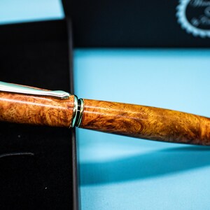 May include: A handcrafted pen with a wood grain barrel and gold-colored accents. The pen features a silver clip and gold-colored tip. The pen is displayed on a black surface with a blue background.