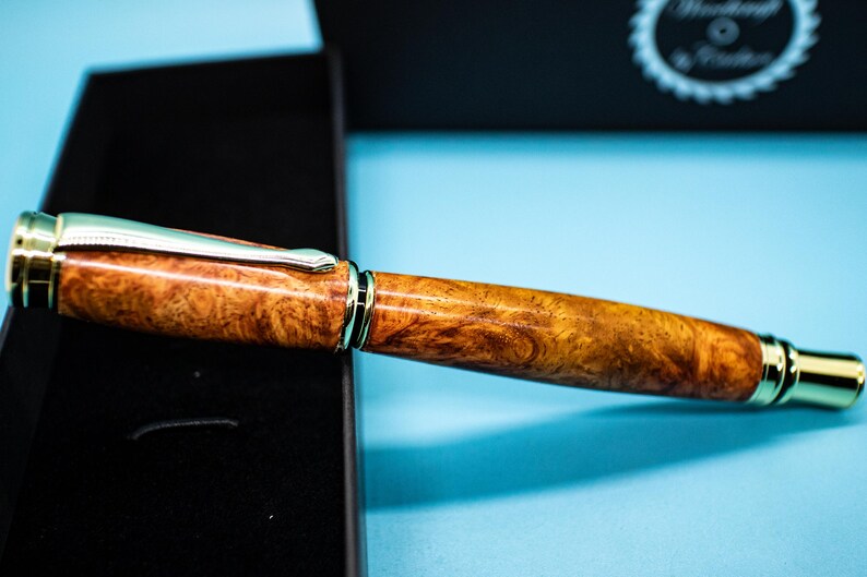 May include: A handcrafted pen with a wood grain barrel and gold-colored accents. The pen features a silver clip and gold-colored tip. The pen is displayed on a black surface with a blue background.