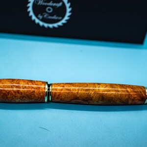 May include: A handcrafted wooden pen with a gold-colored metal trim. The pen features a rich, warm brown burl wood pattern. The pen is displayed on a light blue surface, with a black box in the background that has the text "Woodcraft by Cadence".