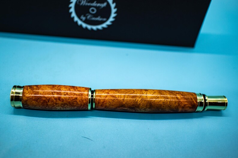 May include: A handcrafted wooden pen with a gold-colored metal trim. The pen features a rich, warm brown burl wood pattern. The pen is displayed on a light blue surface, with a black box in the background that has the text "Woodcraft by Cadence".