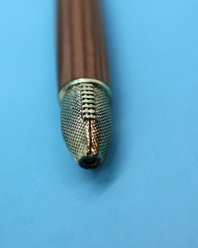 May include: Close-up of a brown wooden pen with a gold-colored tip. The tip has a textured design.