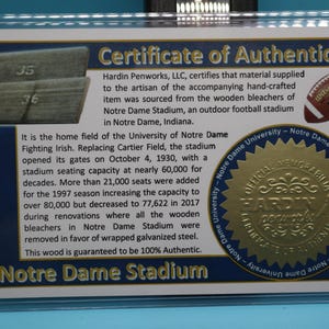 May include: A certificate of authenticity for a piece of wood from the bleachers of Notre Dame Stadium. The certificate is gold and features the Notre Dame Fighting Irish logo. The text on the certificate reads "Hardin Penworks, LLC, certifies that material supplied to the artisan of the accompanying hand-crafted item was sourced from the wooden bleachers of Notre Dame Stadium, an outdoor football stadium in Notre Dame, Indiana. This wood is guaranteed to be 100% Authentic. Notre Dame Stadium."