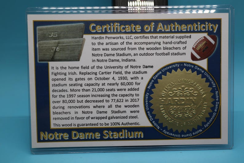 May include: A certificate of authenticity for a piece of wood from the bleachers of Notre Dame Stadium. The certificate is gold and features the Notre Dame Fighting Irish logo. The text on the certificate reads "Hardin Penworks, LLC, certifies that material supplied to the artisan of the accompanying hand-crafted item was sourced from the wooden bleachers of Notre Dame Stadium, an outdoor football stadium in Notre Dame, Indiana. This wood is guaranteed to be 100% Authentic. Notre Dame Stadium."