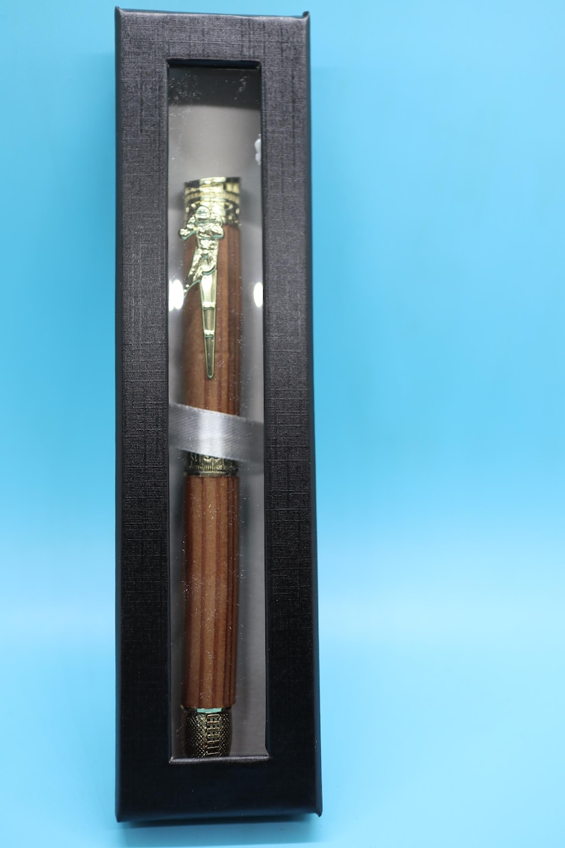 May include: A wooden pen with a gold-colored top and a clear plastic display case. The pen is in a black box with a textured surface.
