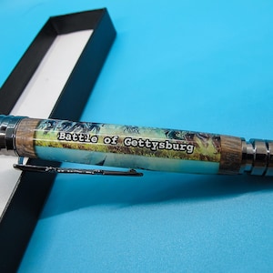 Puede incluir: Un bolígrafo negro con una punta plateada y un cuerpo de madera. El cuerpo tiene una imagen colorida de una escena de batalla y el texto "Battle of Gettysburg".