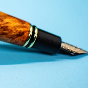 May include: Close-up of a fountain pen with a wooden barrel featuring a unique grain pattern in shades of brown and gold. The pen has a black section with gold accents and a silver nib, set against a light blue background.