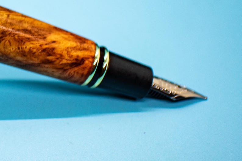 May include: Close-up of a fountain pen with a wooden barrel featuring a unique grain pattern in shades of brown and gold. The pen has a black section with gold accents and a silver nib, set against a light blue background.