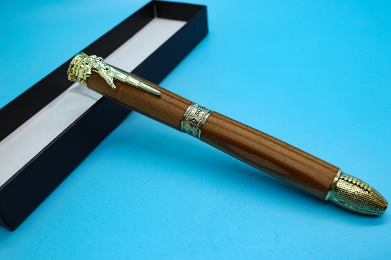 May include: A wooden pen with a gold eagle design on the top and a gold band with numbers around the middle. The pen is in a black box.