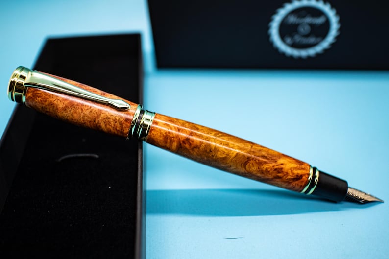 May include: A luxury fountain pen with a burl wood barrel and gold-colored accents. The pen is displayed in a black box against a light blue background. The pen's nib is visible, and the overall design suggests a high-end writing instrument.