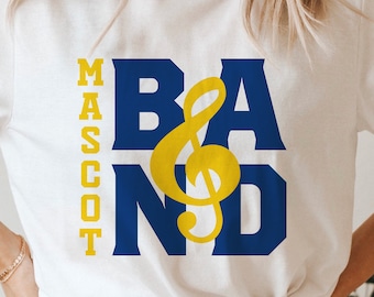 Op de afbeelding: Een wit T-shirt met een blauwe en gele afbeelding waarop "BAND" staat met een sol-sleutel symbool en "MASCOT" verticaal aan de linkerkant. De tekst "FONT INCLUDED for MASCOT and YEAR" staat onderaan de afbeelding.