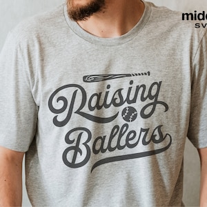 May include: A gray t-shirt with the text "Raising Ballers" in a black script font. A baseball bat and baseball are included in the design.