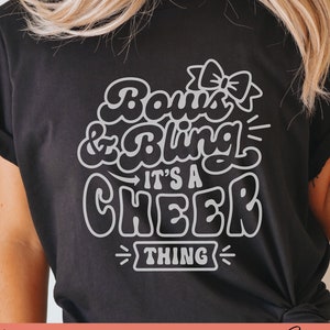 May include: A black t-shirt with a white design that says "Bows & Bling It's a Cheer Thing".