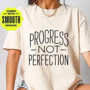 May include: A white t-shirt with the text "PROGRESS - NOT - PERFECTION" printed in black. The text has a distressed, vintage look. The shirt is being modeled by a person. The image also includes a yellow circle with the text "COMES WITH SMOOTH VERSION".