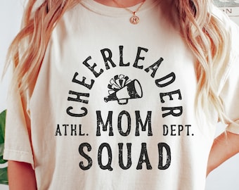 May include: A white t-shirt with a black graphic design that says "Cheerleader Mom Squad" with a megaphone and pom-poms in the center.