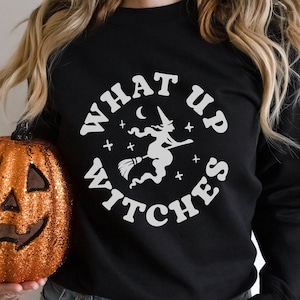 Puede incluir: Una sudadera negra con un diseño gráfico blanco que dice "What Up Witches" con una bruja volando en una escoba dentro de un círculo.