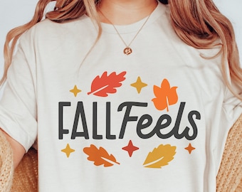 May include: A white t-shirt with the text "FALL Feels" in black lettering surrounded by orange, red, and yellow leaves and gold stars.