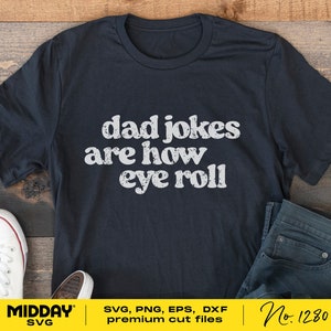 May include: A black t-shirt with white text that reads "dad jokes are how eye roll". The shirt is lying on a wooden surface with a pair of white trainers and blue jeans.