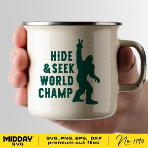 May include: A white enamel mug with a green silhouette of Bigfoot giving the peace sign. The text "HIDE & SEEK WORLD CHAMP" is printed in green on the mug.