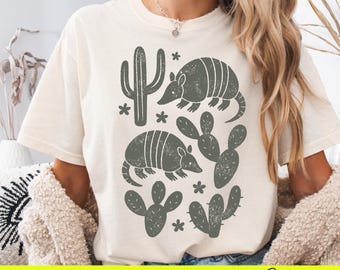 May include: A white t-shirt with a graphic design featuring armadillos, cacti, and a stylized cactus. The design is in shades of gray and brown.