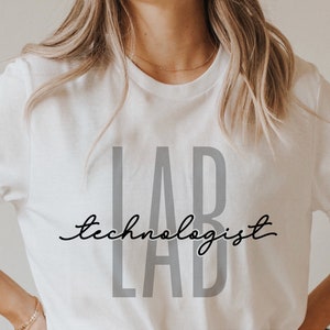 May include: A white t-shirt with the words "LAB" in gray block letters and "technologist" in a cursive font. The word "LAB" is repeated below the word "technologist". The shirt has short sleeves.