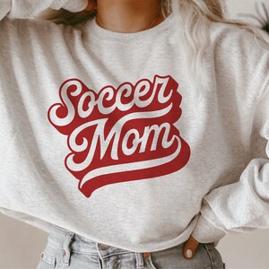 K&ouml;nnte beinhalten: Ein grauer Pullover mit einer roten und wei&szlig;en Grafik, auf der in einer Retro-Schrift "Soccer Mom" steht.