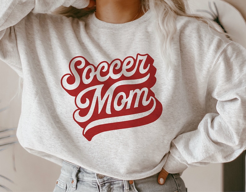 K&ouml;nnte beinhalten: Ein grauer Pullover mit einer roten und wei&szlig;en Grafik, auf der in einer Retro-Schrift "Soccer Mom" steht.