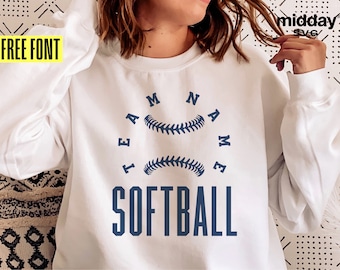 以下が含まれることがあります： 白のスウェットシャツ。ネイビーブルーのブロック体で「SOFTBALL」の文字。その上に、ネイビーブルーの野球ボールが2つあり、その周りに「TEAM NAME」の文字が半円状に配置されています。「FREE FONT」の文字が左上の黄色のボックスにあります。