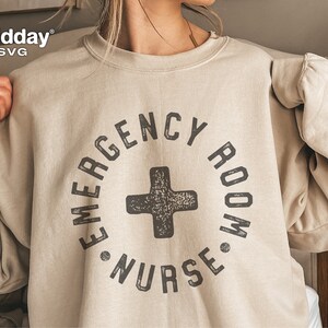 Peut inclure: Un sweat-shirt beige avec un imprim&eacute; graphique noir qui dit "Emergency Room Nurse" dans un cercle avec un signe plus noir au centre.