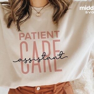 May include: A beige sweatshirt with the text "PATIENT CARE assistant" in black and pink lettering.