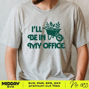 May include: A light grey t-shirt featuring the text "I'LL BE IN MY OFFICE" in dark green. The design includes a wheelbarrow filled with plants. This is a casual wear item.