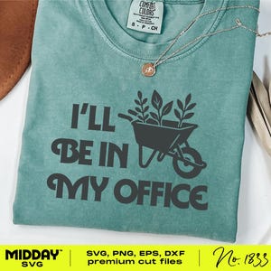 May include: A teal t-shirt with the text "I'll Be In My Office" in dark grey. A graphic of a wheelbarrow filled with plants is above the words. A gold necklace with a small pendant is visible. The shirt has a Comfort Colors label.