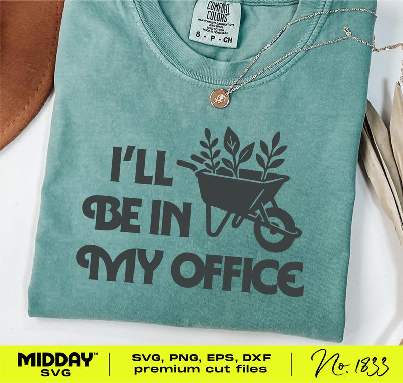 May include: A teal t-shirt with the text "I'll Be In My Office" in dark grey. A graphic of a wheelbarrow filled with plants is above the words. A gold necklace with a small pendant is visible. The shirt has a Comfort Colors label.