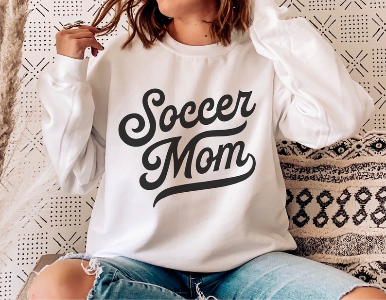 Może przedstawiać: Biała bluza z czarnym napisem "Soccer Mom" w retro czcionce.