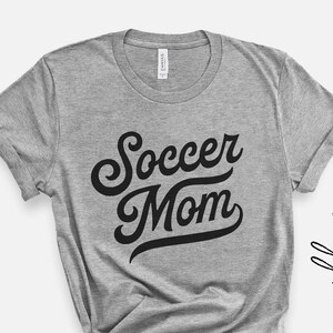 Może przedstawiać: Szara koszulka z czarnym napisem "Soccer Mom" w retro czcionce.