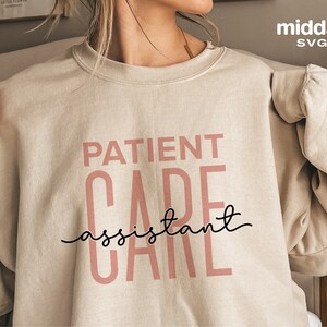 May include: A beige sweatshirt with the text "PATIENT CARE assistant CARE" printed in pink letters. The text "assistant" is written in black cursive.