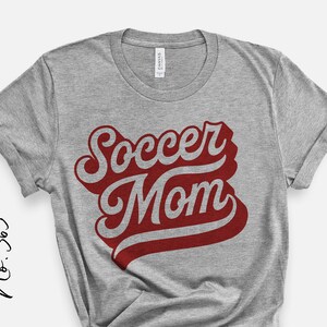 K&ouml;nnte beinhalten: Graues T-Shirt mit dem Text "Soccer Mom" in roten und wei&szlig;en Buchstaben.