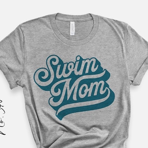 Puede incluir: Camiseta gris con el texto "Swim Mom" en letras azul turquesa. El texto está en una fuente retro y estilizada.