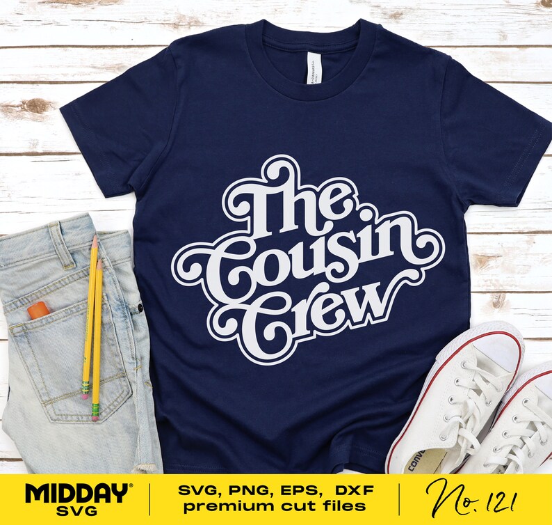 May include: A navy blue t-shirt with white text that reads "The Cousin Crew" in a retro font style. The shirt is laid flat on a wooden surface with a pair of jeans and a pair of white sneakers.
