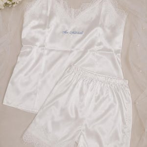 May include: A white satin camisole and shorts pyjama set featuring delicate lace trim along the neckline and hem. The camisole has thin spaghetti straps and the text Mrs Mitchell embroidered in light blue thread on the front chest area.