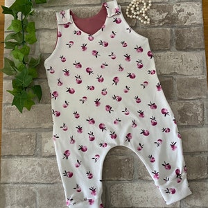 May include: White baby jumpsuit with a pink and white apple print. The jumpsuit has a pink lining and is sleeveless. The text "Mini Makes by Julia" is visible on a wooden sign in the background.