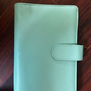 May include: A light teal-colored planner with a leather-like texture. The planner has a rectangular shape with rounded corners and a flap closure. The stitching is visible around the edges. The planner is closed and resting on a wooden surface.