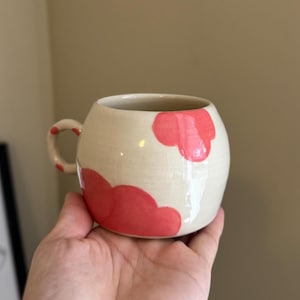 May include: Cream ceramic mug with a rounded shape and a small handle. The mug is decorated with bright red cloud-like shapes. The mug is held in a hand.