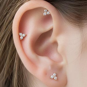 May include: Close-up of an ear with three small, gold-coloured earrings. Each earring has a cluster of three clear, sparkling stones, creating a delicate design. The earrings are set against the skin, highlighting the jewellery.