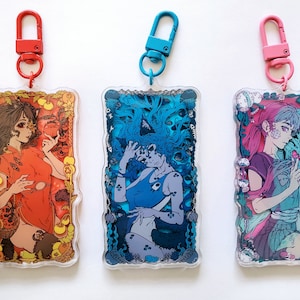 May include: Three rectangular acrylic keychains with colorful illustrations of anime-style figures. Each keychain has a different color clip: red, blue, and pink. The illustrations feature detailed artwork with various color schemes and themes.